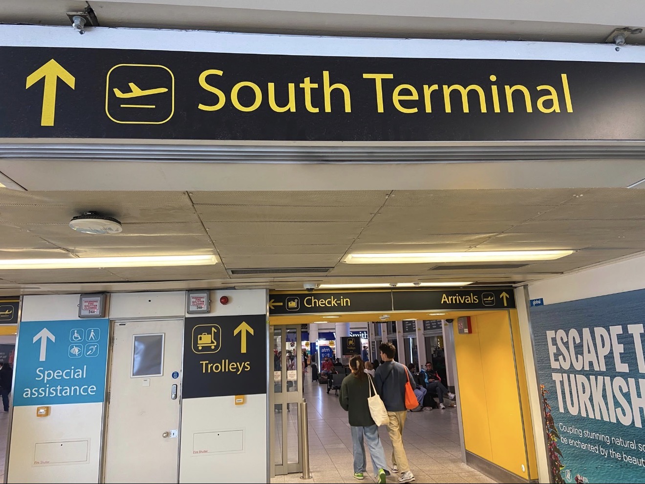 Yellow-on-black South Terminal sign at Gatwick Airport above a walkway signed for check-in and arrivals