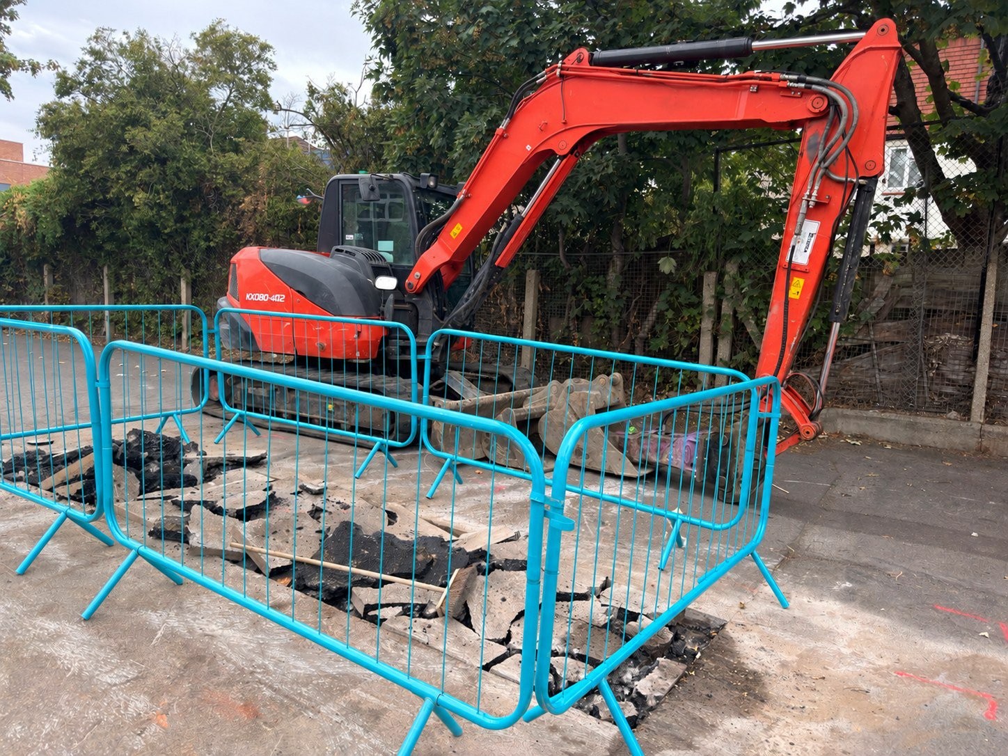 Orange tracked excavator beside a fenced trial pit where the hard surface has been broken up, with trees behind
